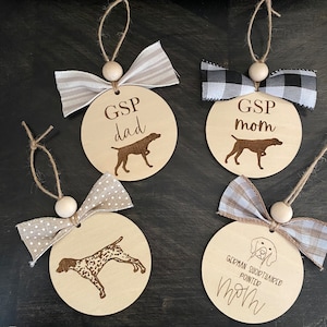 May include: Four round wooden ornaments with brown twine and a bow. Each ornament features a silhouette of a German Shorthaired Pointer dog and text. The ornaments read "GSP dad", "GSP mom", "German Shorthaired Pointer mom", and "GSP mom".