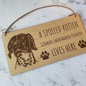 Könnte beinhalten: Holzschild mit dem Text „A SPOILED ROTTEN German Shorthaired Pointer LIVES HERE“. Das Schild zeigt ein detailliertes eingraviertes Bild eines Hundekopfes und zwei Pfotenabdrücke. Eine Lederschnur ist zum Aufhängen befestigt.