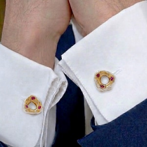 Puede incluir: Un par de gemelos de oro con una forma circular orgánica e irregular, cada uno con tres pequeñas piedras preciosas redondas de color rojo intenso incrustadas en la superficie metálica texturizada, sujetos a los puños blancos de una camisa.