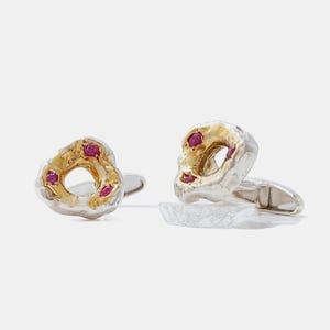 Puede incluir: Un par de gemelos con un diseño orgánico e irregular, con un exterior de plata texturizada y un centro en tono dorado. Cada pieza está decorada con tres pequeños rubíes redondos de color rosa vibrante incrustados en la superficie dorada.
