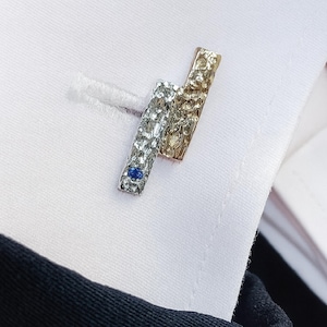 Peut inclure: Gros plan sur une paire de boutons de manchette. Un bouton de manchette est argenté avec une surface texturée et une petite pierre précieuse bleue. L'autre est doré avec une texture martelée. Sur une chemise blanche.
