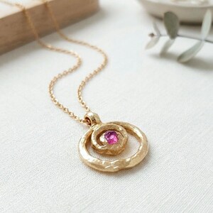 Könnte beinhalten: Goldfarbene Halskette mit Anhänger und einem pinkfarbenen Edelstein. Der Anhänger hat ein Spiraldesign in einem größeren Kreis, das an einer zarten Kette hängt. Die Halskette wird auf einer weißen Oberfläche präsentiert.