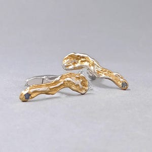 Könnte beinhalten: Ein Paar goldfarbene Manschettenknöpfe mit einem strukturierten, organischen Design. Die Manschettenknöpfe haben eine silberne Rückseite und einen kleinen, dunkel gefärbten Stein an jedem Ende. Das Design ist abstrakt und modern.