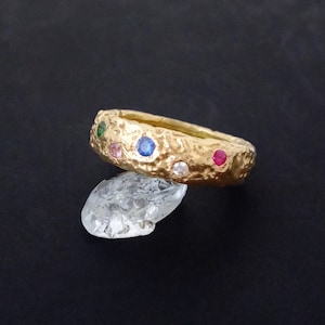 May include: Gold ring with a textured band and colourful gemstones. The ring is set with a variety of small, round gemstones in shades of pink, blue, green, and red.