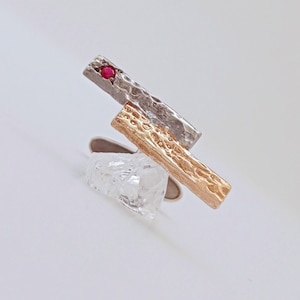 May include: A modern ring featuring a clear, irregular-shaped stone and two textured metal bars. One bar is silver with a red gemstone, the other is gold. The ring is set against a white background.