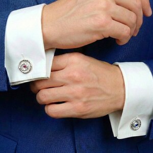 May include: Close-up of a person wearing a blue suit and white dress shirt with silver cufflinks. The cufflinks have a circular design with a red gemstone in the center, surrounded by a silver border. The shirt cuffs are neatly folded.