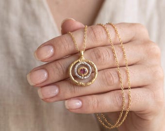 May include: A gold-toned necklace featuring a pendant with two concentric circular rings. The outer ring has a textured, hammered finish, while the inner ring surrounds a small, oval-shaped, deep pink gemstone set in a gold-toned bezel.