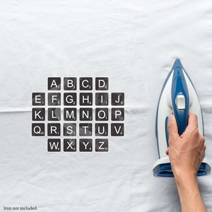 May include: A white fabric surface with a set of black alphabet keys and a blue and white iron. The keys are labeled with letters and numbers. The iron is held by a hand.
