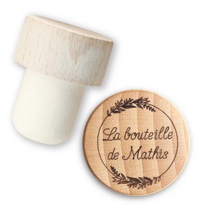 May include: A wooden bottle stopper with a round, engraved top. The top features a wreath of leaves and the text "La bouteille de Mathis".