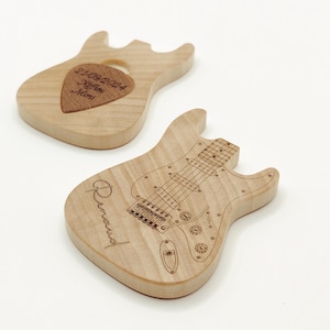 Può includere: Due ornamenti in legno a forma di chitarra. Uno è una chitarra a forma di plettro con il testo "21.09.2024  Kiffen Mimi" inciso su di essa. L'altro è una chitarra a forma completa con il testo "Renaud" inciso su di essa.