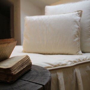 May include: An open book lies on a wooden stump in front of a white bed with two white pillows. The bed has a white ruffle at the bottom.