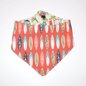 May include: A triangular pet bandana featuring a vibrant coral-red background patterned with rows of stylized surfboard shapes in various colors and textures. The top fold displays a cream-colored fabric with beach huts and a bicycle, secured by a metal snap.