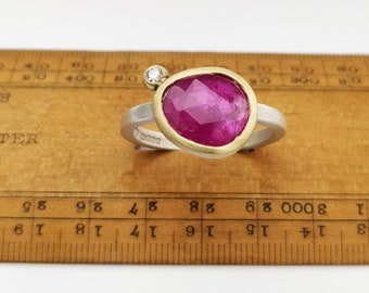 May include: A ring with a pink ruby gemstone set in a gold bezel. The ring is made of silver and gold and has a small diamond accent.