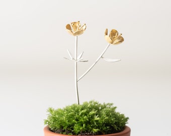 Puede incluir: Una pequeña escultura de flores de metal dorado con dos flores y tallos plateados, colocada en una maceta de musgo verde.