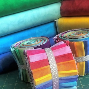 May include: Two rolls of rainbow fabric scraps, each tied with a white ribbon with white polka dots. The fabric scraps are arranged in a rainbow pattern, with red, orange, yellow, green, blue, and purple. The fabric scraps are on a green cutting mat.