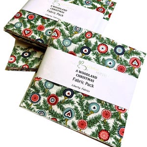 May include: Fabric packs with a woodland Christmas pattern. The fabric displays a white backdrop with green fir branches and red, blue, and teal ornaments. Each pack is wrapped with a white band that reads "A WOODLAND CHRISTMAS Fabric Pack".
