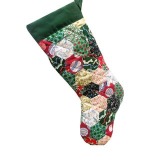 以下が含まれることがあります： パッチワークデザインの手作りクリスマスストッキング。上部は無地の濃い緑色で、ボディは赤、緑、白、金色のさまざまな柄の生地を使用しています。ストッキングは吊るすように設計されています。