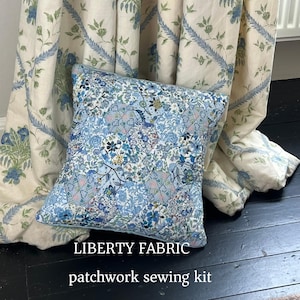 May include: A blue floral patchwork cushion with a matching curtain backdrop. The cushion features a detailed pattern in blue, white, and yellow. The text "LIBERTY FABRIC" and "patchwork sewing kit" are displayed below the cushion.