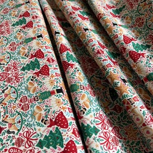 May include: A white fabric featuring a repeating Christmas pattern of nutcrackers, ballerinas, green pine trees, gold bells, red bows, and heart-shaped ornaments. The festive design includes small stars and sprigs of holly scattered across the material.