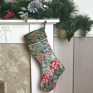 Peut inclure: Une chaussette de Noël matelassée avec un motif patchwork à motifs floraux rouges, verts et blancs. Le revers est vert foncé avec un imprimé floral. La chaussette est suspendue à une cheminée blanche ornée de verdure et d'une boule dorée.