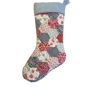 以下が含まれることがあります： パッチワークデザインのキルティングクリスマスストッキング。ストッキングは、ライトブルーのカフとループ、赤いトリムが特徴です。ストッキングの本体は、赤、白、グレーのさまざまな模様の生地で作られています。