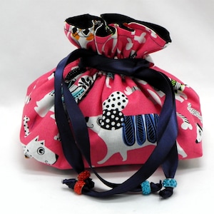 May include: Pink drawstring bag with a white and black dog print. The bag has a navy blue ribbon with red and blue beads.
