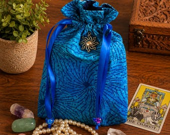 Puede incluir: Una bolsa de tela azul con cordón, con un patrón de estallido de estrellas azul oscuro, un dije de sol metálico y dos cintas de satén azul con cuentas. La bolsa está rodeada por un collar de perlas blancas, piedras pulidas y una carta del tarot.