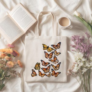 Puede incluir: Un bolso de tela color crema con un dise&ntilde;o estampado de varias mariposas monarca en tonos naranja, negro y blanco. Las mariposas est&aacute;n dispuestas en un grupo en la parte frontal del bolso, el cual cuenta con dos asas de tela.