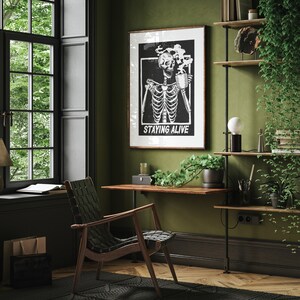 May include: A black and white illustration of a skeleton drinking from a mug with the text "Staying Alive" on a black background. The illustration is framed and hanging on a green wall in a home office.