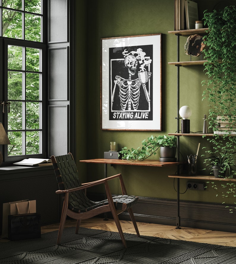 May include: A black and white illustration of a skeleton drinking from a mug with the text "Staying Alive" on a black background. The illustration is framed and hanging on a green wall in a home office.
