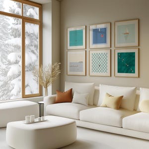 K&ouml;nnte beinhalten: Ein wei&szlig;es Sofa mit braunen und cremefarbenen Kissen steht vor einer Wand mit einer Galerie gerahmter Kunstwerke. Die Kunstwerke zeigen abstrakte Designs in Blaut&ouml;nen, Gr&uuml;nt&ouml;nen und Wei&szlig;. Ein gro&szlig;es Fenster hinter dem Sofa zeigt eine verschneite Winterlandschaft.