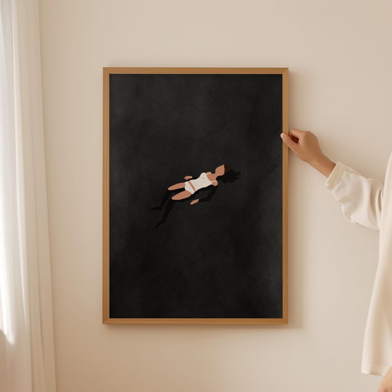 May include: A rectangular wall art print in a light wood frame featuring a dark charcoal background with a centered illustration of a figure in a white swimsuit floating horizontally. The figure has dark hair and tan skin, depicted in a simplified style.