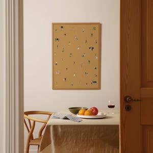 May include: A framed art print featuring a mustard-colored background with numerous tiny figures scattered across it. The print is displayed on a wall above a table set with fruit, a bowl, and a glass of wine. The frame is a light brown color.