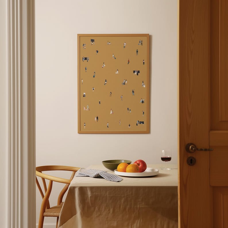 May include: A framed art print featuring a mustard-colored background with numerous tiny figures scattered across it. The print is displayed on a wall above a table set with fruit, a bowl, and a glass of wine. The frame is a light brown color.