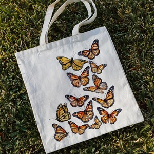 Puede incluir: Un bolso de tela de color crema con un dise&ntilde;o estampado de numerosos ejemplares de mariposa monarca en tonos naranja, negro y blanco. Las mariposas est&aacute;n dispuestas en un patr&oacute;n en cascada sobre la parte frontal del bolso rectangular.