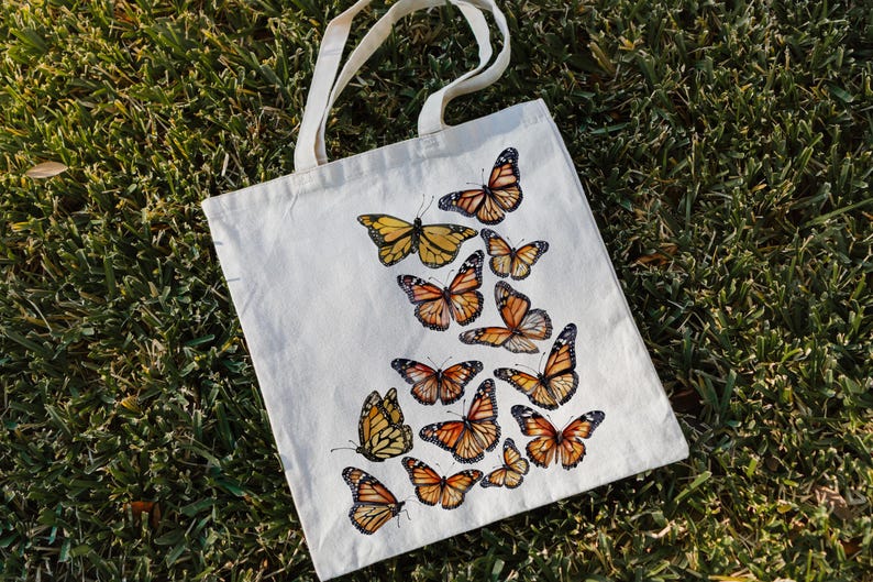Puede incluir: Un bolso de tela de color crema con un dise&ntilde;o estampado de numerosos ejemplares de mariposa monarca en tonos naranja, negro y blanco. Las mariposas est&aacute;n dispuestas en un patr&oacute;n en cascada sobre la parte frontal del bolso rectangular.