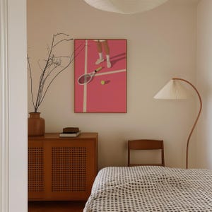May include: A pink tennis court with a tennis racket and a tennis ball on the court. The image is framed and hanging on a wall in a bedroom with a bed, a lamp, and a dresser.
