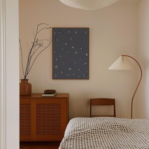 May include: A rectangular wall art print featuring a dark slate blue background with scattered small, abstract, multi-colored figures and shapes, enclosed in a thin wooden frame and mounted on a plain, light-colored wall above a wooden cabinet.