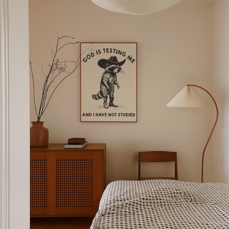 Puede incluir: Un cuadro enmarcado con un mapache con sombrero de vaquero y botas. El texto en el cuadro dice "GOD IS TESTING ME" y "AND I HAVE NOT STUDIED". El cuadro est&aacute; en un dormitorio con un mueble de madera y una l&aacute;mpara de pie.