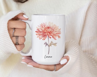 Puede incluir: Una taza de cerámica blanca con una ilustración estilo acuarela de un crisantemo en tonos melocotón y rosa con hojas verdes en el tallo. El nombre Emma está impreso en una fuente cursiva negra debajo del diseño floral en la taza.