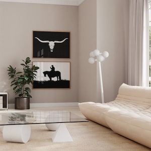 May include: A living room interior with a white sofa, a glass coffee table, and two black and white framed prints on the wall. The prints depict a cowboy riding a horse and a longhorn skull.