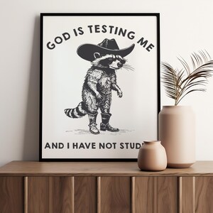 Puede incluir: L&aacute;mina art&iacute;stica en blanco y negro que representa un mapache con sombrero y botas de vaquero. El texto superior dice "GOD IS TESTING ME" y debajo "AND I HAVE NOT STUD". La impresi&oacute;n se muestra en un marco negro.