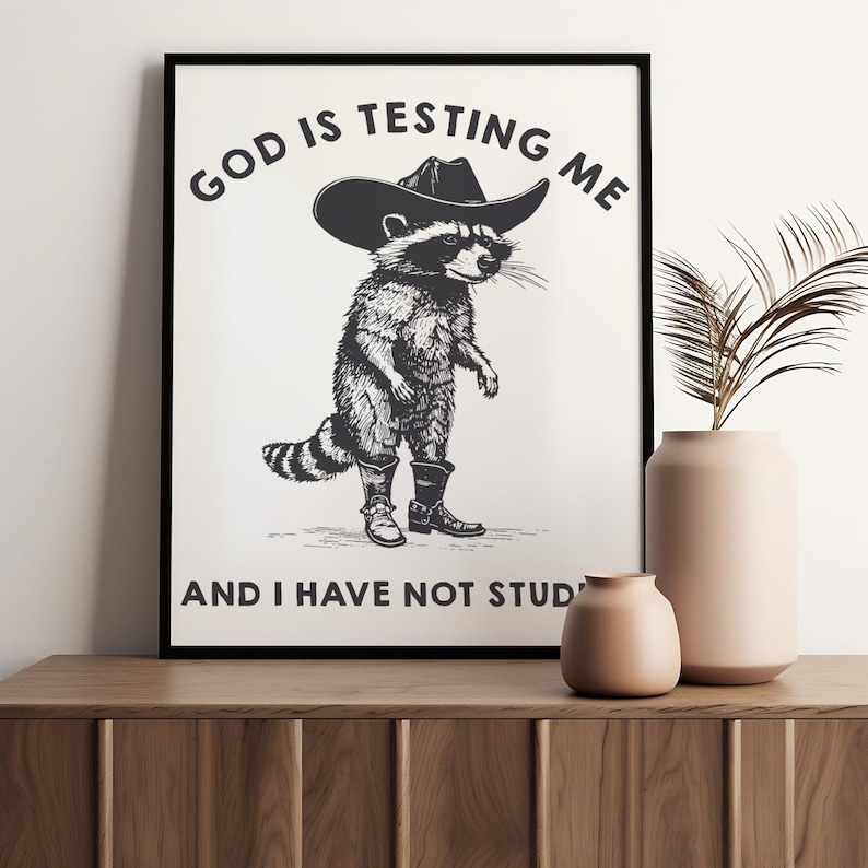 Puede incluir: L&aacute;mina art&iacute;stica en blanco y negro que representa un mapache con sombrero y botas de vaquero. El texto superior dice "GOD IS TESTING ME" y debajo "AND I HAVE NOT STUD". La impresi&oacute;n se muestra en un marco negro.