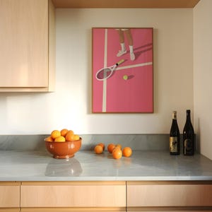 May include: A kitchen counter with a bowl of oranges, two bottles of wine, and a framed print of a tennis player on a pink court.