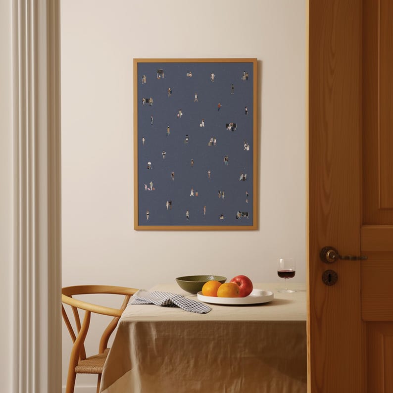 May include: A rectangular wall art print in a light wood frame featuring a dark blue background with a scattered pattern of tiny, diverse human figures. The print is displayed on a plain wall above a dining table with a beige tablecloth and fruit.