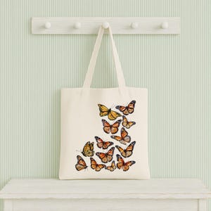 Puede incluir: Un bolso de tela color crema con un dise&ntilde;o estampado de varias mariposas monarca en tonos naranja, negro y blanco. Las mariposas est&aacute;n dispuestas en un grupo en la parte inferior derecha del bolso, el cual cuenta con dos asas.