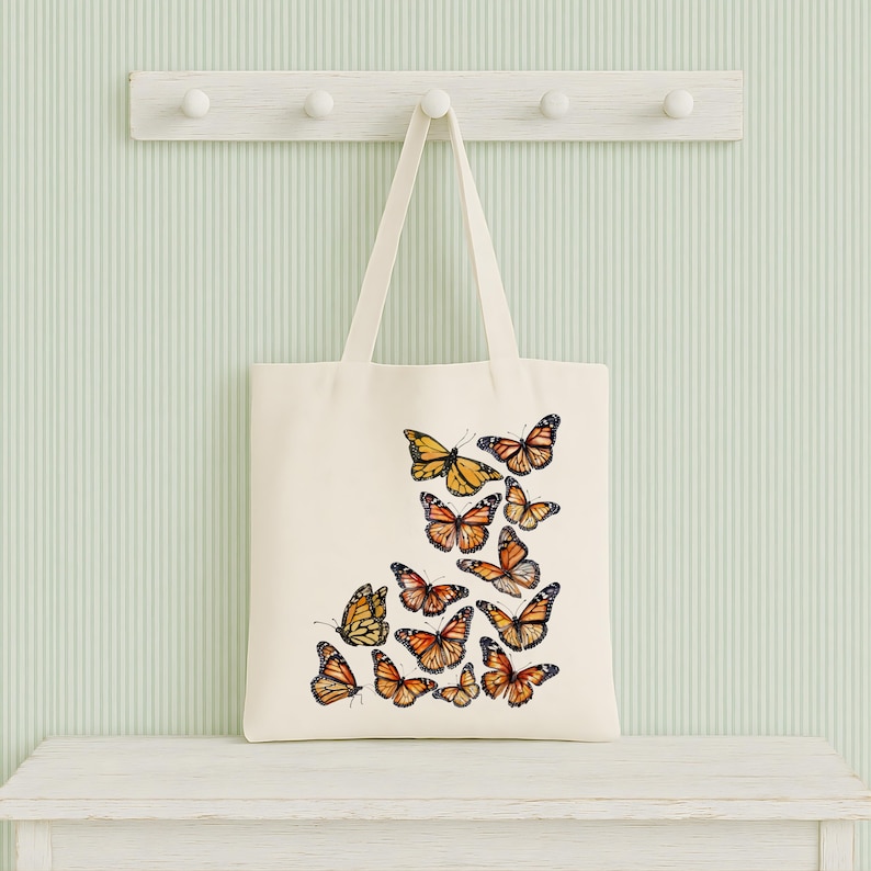 Puede incluir: Un bolso de tela color crema con un dise&ntilde;o estampado de varias mariposas monarca en tonos naranja, negro y blanco. Las mariposas est&aacute;n dispuestas en un grupo en la parte inferior derecha del bolso, el cual cuenta con dos asas.