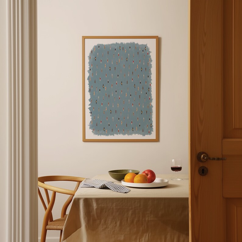 May include: A framed print of a blue background with a pattern of small, colorful figures. The print is hanging on a white wall above a table with a white tablecloth, a bowl of fruit, and a glass of wine.