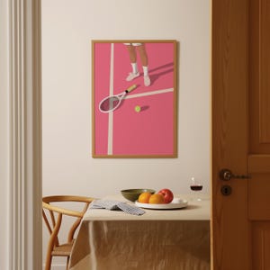 May include: A framed illustration of a tennis player on a pink court with a tennis racket and ball. The illustration is hanging on a white wall above a wooden table with a bowl of fruit.