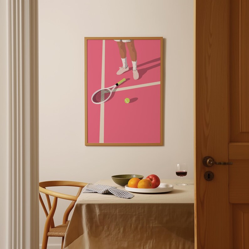 May include: A framed illustration of a tennis player on a pink court with a tennis racket and ball. The illustration is hanging on a white wall above a wooden table with a bowl of fruit.